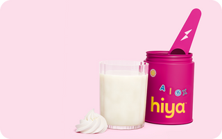 Pink Hiya kids' nutrient powder container with spoon, glass of milk, and whipped cream on pink background
