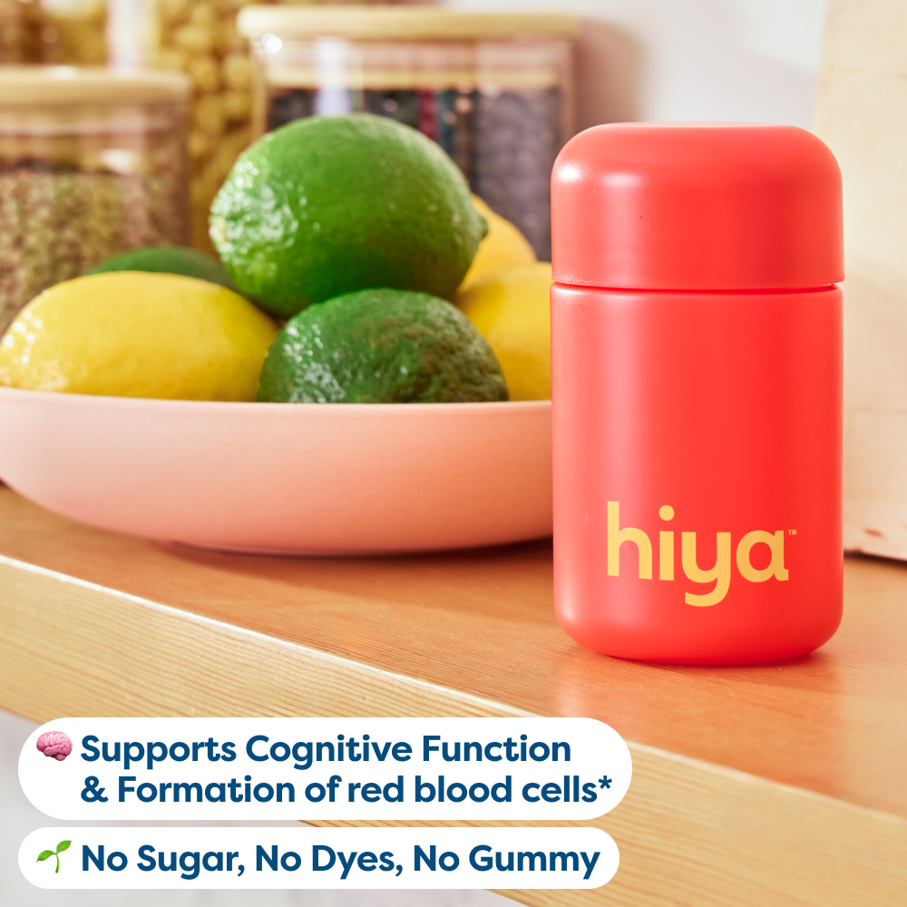 A bright red cylindrical bottle with the brand name 'hiya' in yellow on the front, placed on a wooden surface next to a bowl of lemons and limes. Two white text banners at the bottom read 'Supports Cognitive Function & Formation of red blood cells*' and 'No Sugar, No Dyes, No Gummy'.