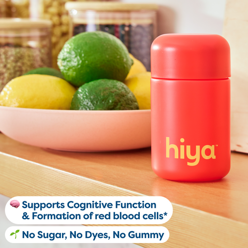 A bright red cylindrical bottle with the brand name 'hiya' in yellow on the front, placed on a wooden surface next to a bowl of lemons and limes. Two white text banners at the bottom read 'Supports Cognitive Function & Formation of red blood cells*' and 'No Sugar, No Dyes, No Gummy'.
