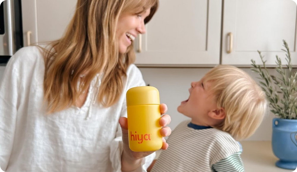 Mom holding yellow Hiya vitamin bottle with smiling child in kitchen