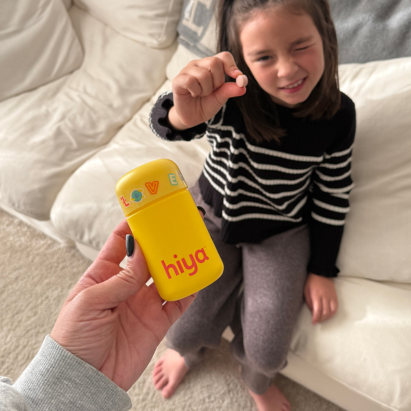 Child holding vitamin tablet with yellow Hiya Health bottle, promoting kids' nutrition and healthy choices