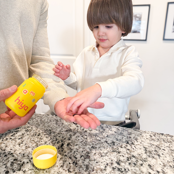 Child taking vitamin tablet from parent’s hand, yellow bottle labeled hiya on granite counter